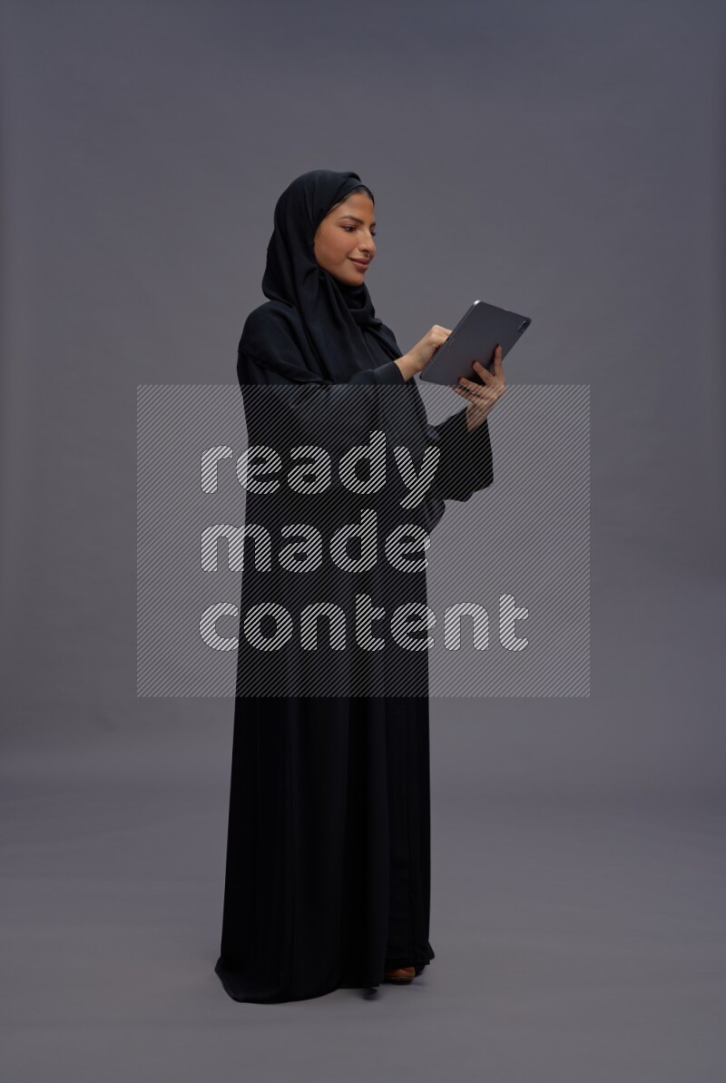 Saudi woman wearing Abaya standing working on tablet on gray background