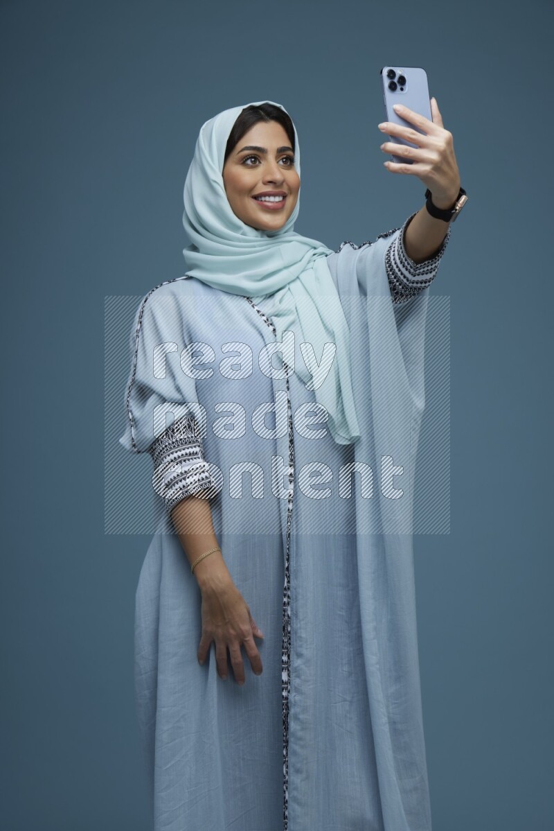 A Saudi woman Taking a Selfie on a blue background wearing a blue Abaya with hijab