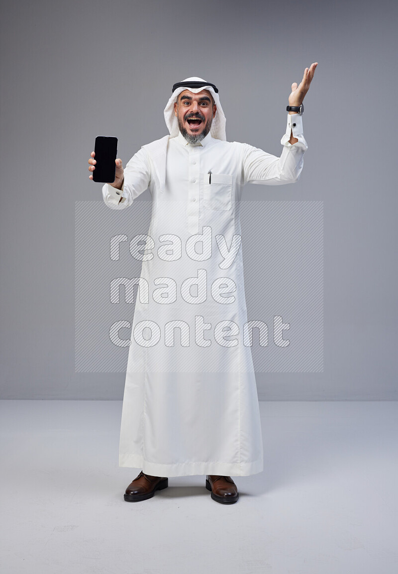 Saudi man Wearing Thob and white Shomag standing showing phone to camera on Gray background