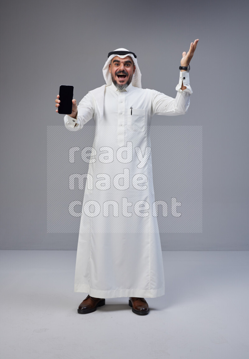 Saudi man Wearing Thob and white Shomag standing showing phone to camera on Gray background