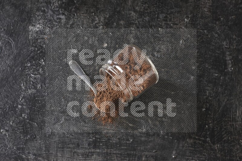 وعاء زجاجي مقلوب وملعقة معدنية ممتلئان ببودرة القرنفل مع حبوب القرنفل المتناثرة على أرضية سوداء