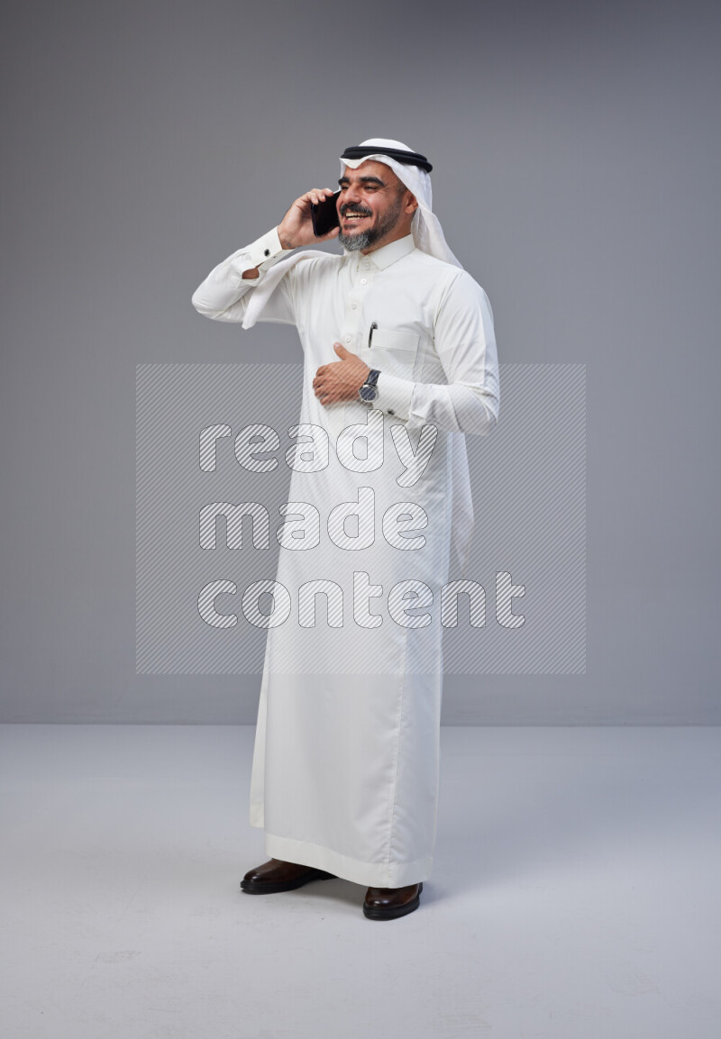 Saudi man Wearing Thob and white Shomag standing talking on phone on Gray background