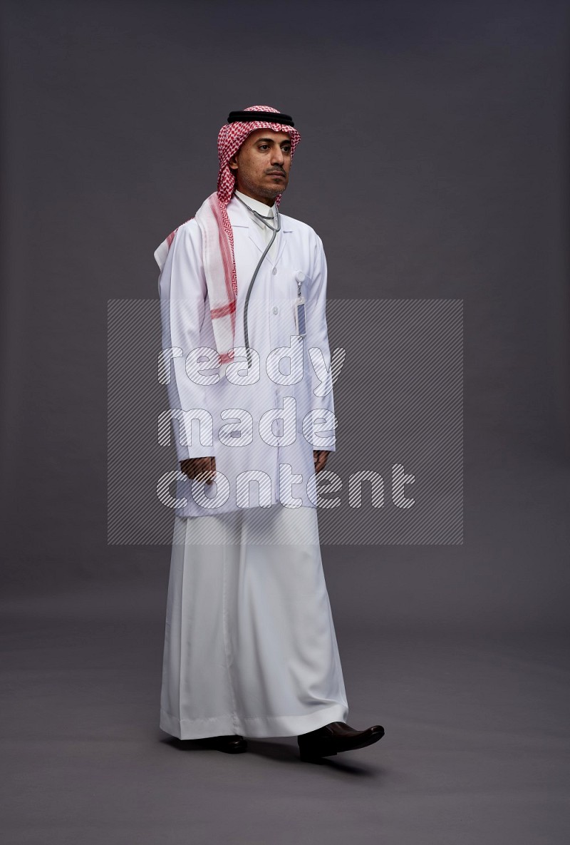 Saudi man wearing thob with lab coat and shomag with pocket employee badge with stethoscope standing interacting with the camera on gray background