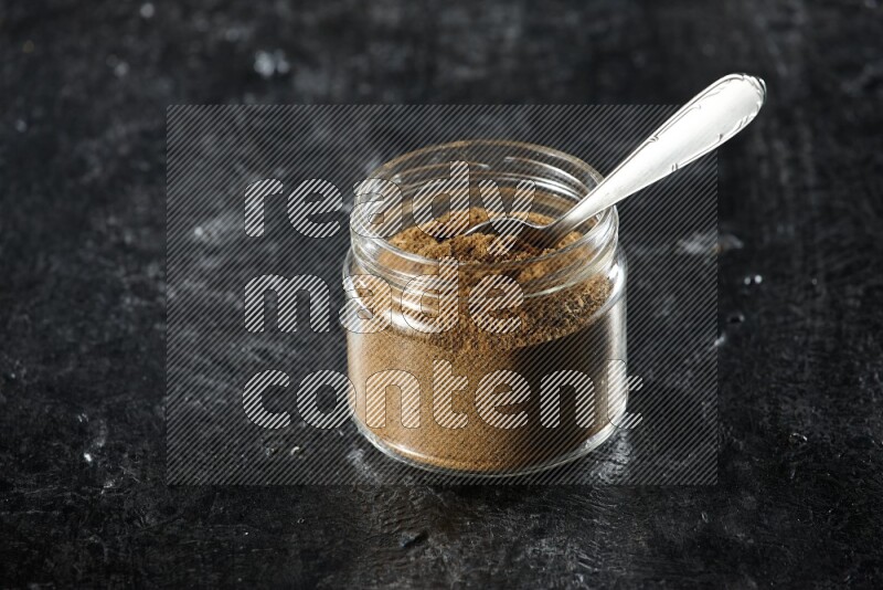 A glass jar and a metal spoon full of cumin powder on a textured black flooring