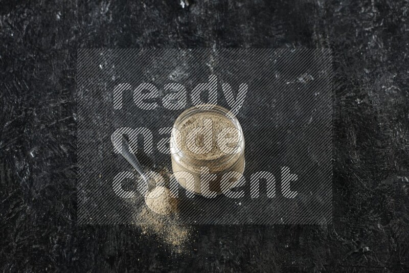 A glass jar and metal spoon full of cardamom powder on textured black flooring