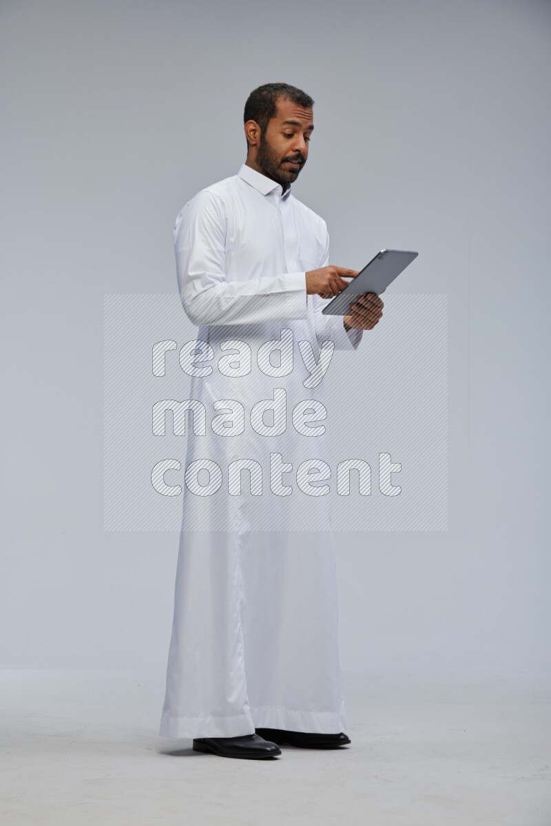 Saudi man wearing Thob standing working on tablet on Gray background