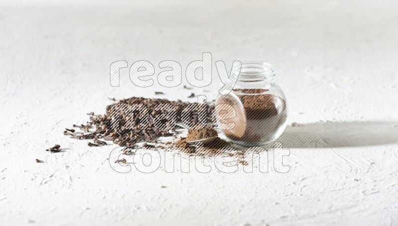 A glass spice jar and a metal spoon full of cloves powder and cloves spread on textured white flooring