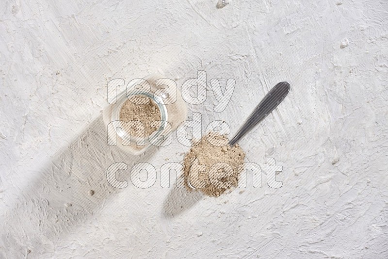 A glass spice jar full of garlic powder with metal spoon on a textured white flooring in different angles