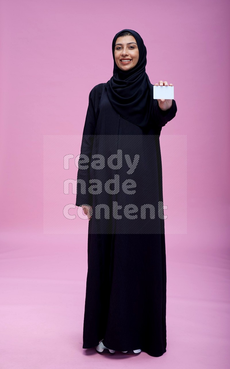 Saudi woman wearing Abaya standing holding ATM card on pink background