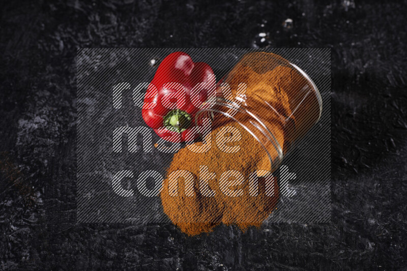 A glass jar full of ground paprika powder flipped with some spilling powder on black background