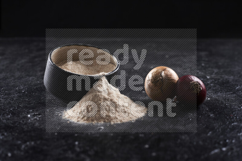 A black pottery bowl full of onion powder with fallen powder from it on black background