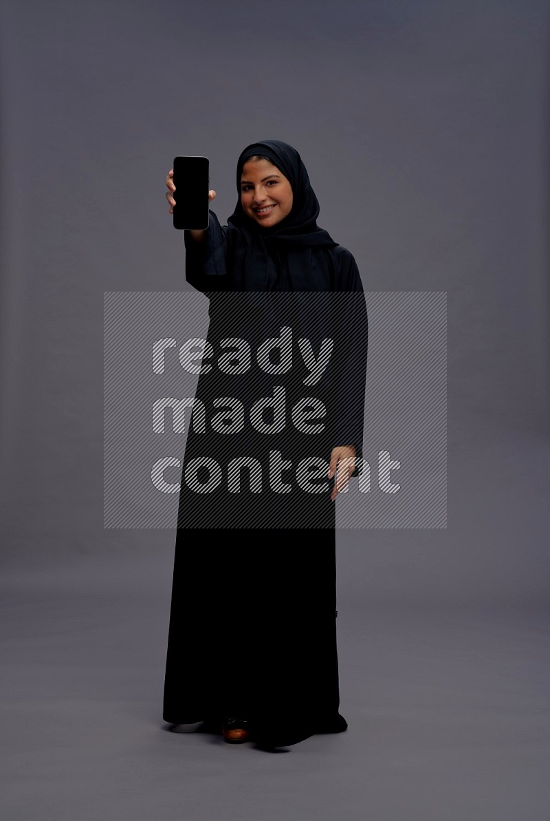 Saudi woman wearing Abaya standing showing phone to camera on gray background