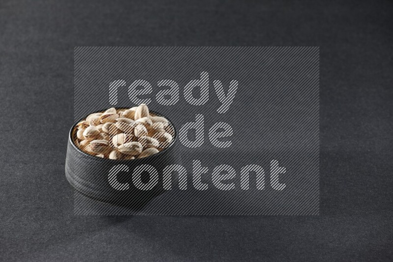 A black pottery bowl full of pistachios on a black background in different angles