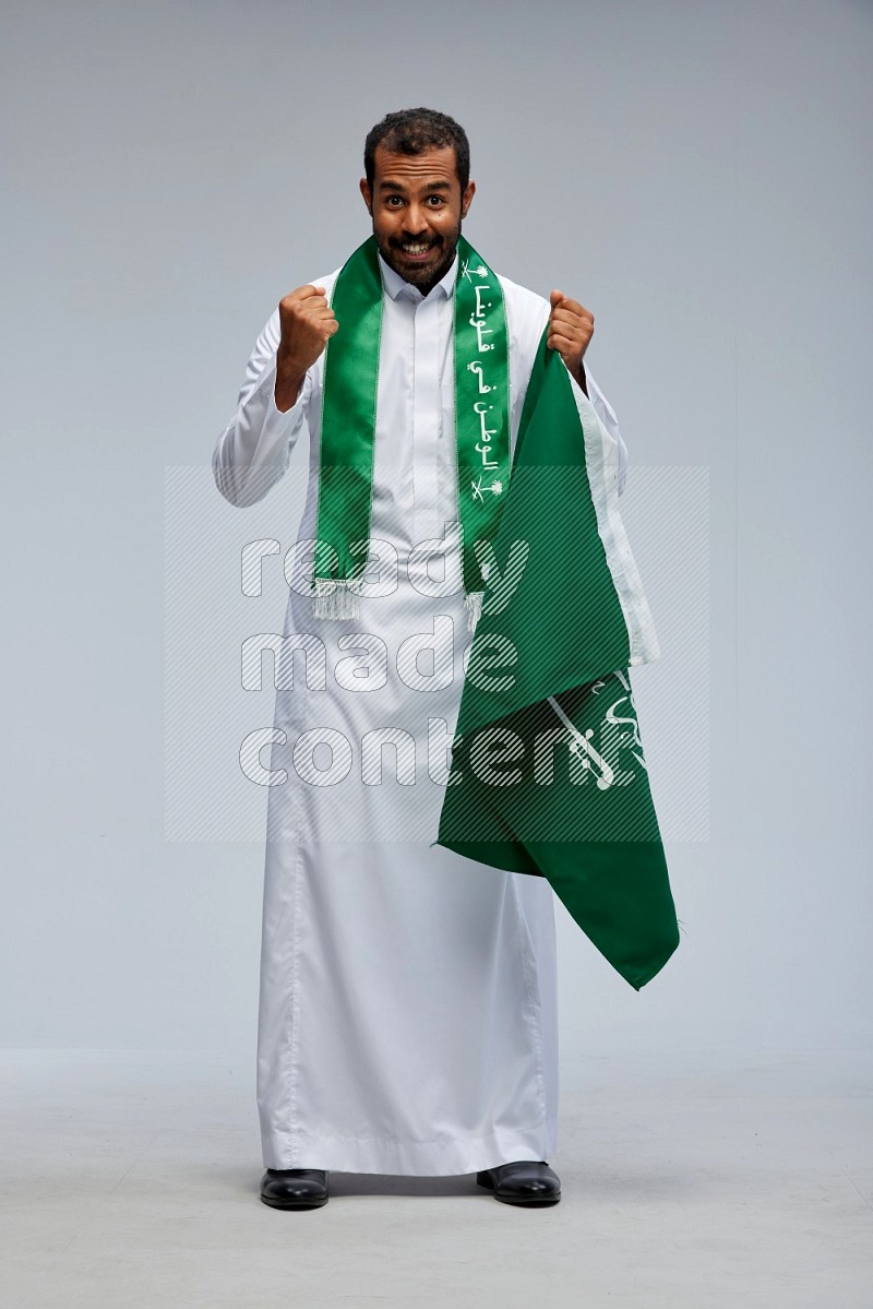 Saudi man wearing Thob standing holding Saudi flag on Gray background