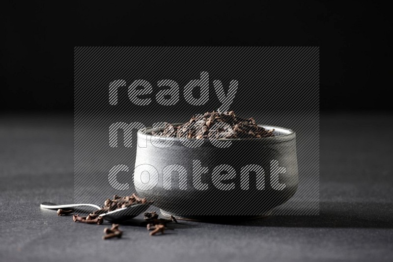 A black pottery bowl full of cloves and a metal spoon next to it on a black flooring