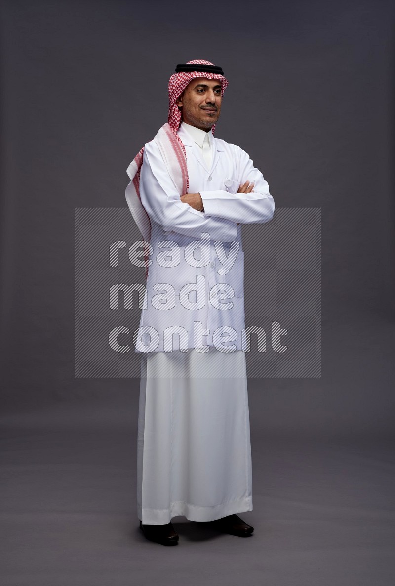 Saudi man wearing thob with lab coat and shomag with pocket employee badge standing with crossed arms on gray background