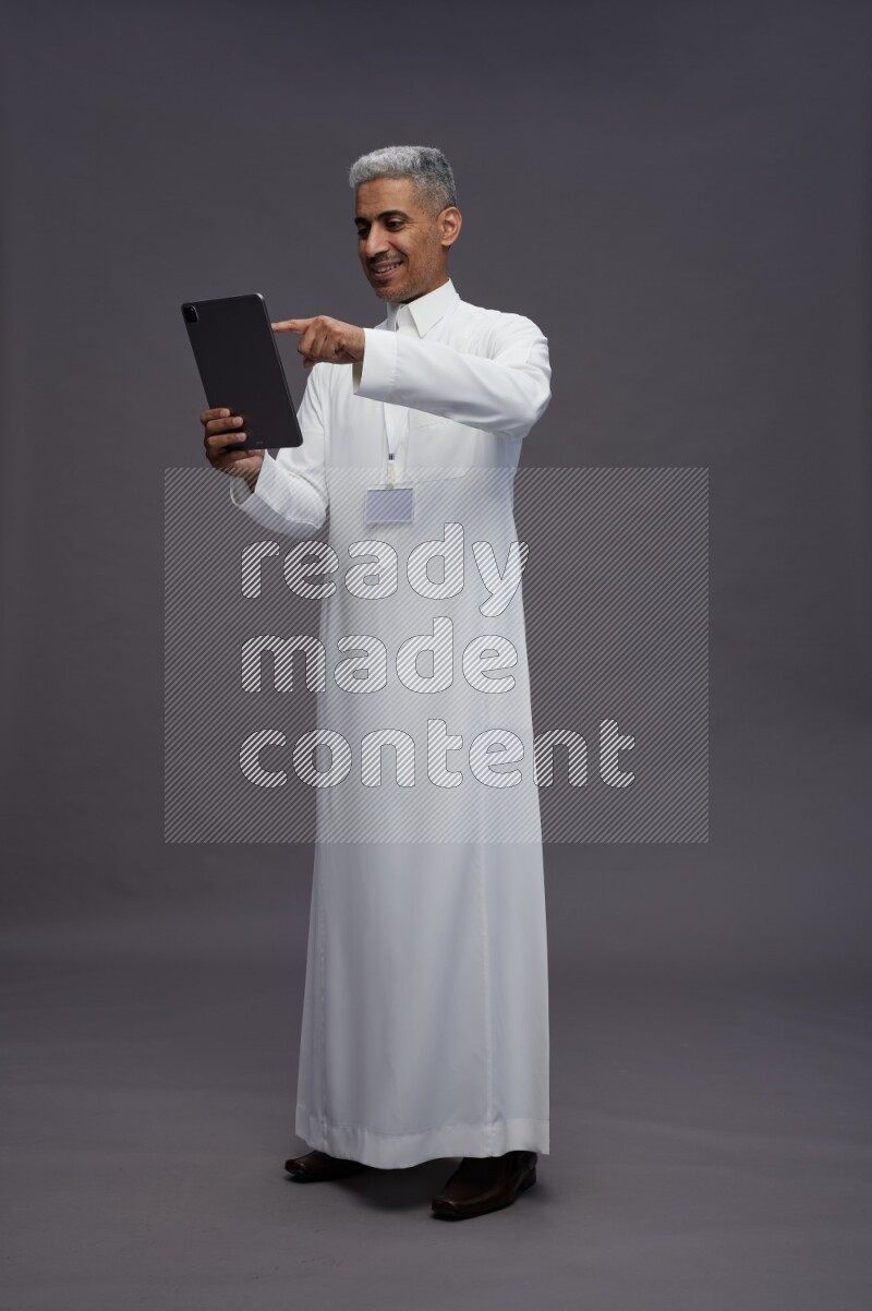 Saudi man wearing thob with neck strap employee badge standing working on tablet on gray background