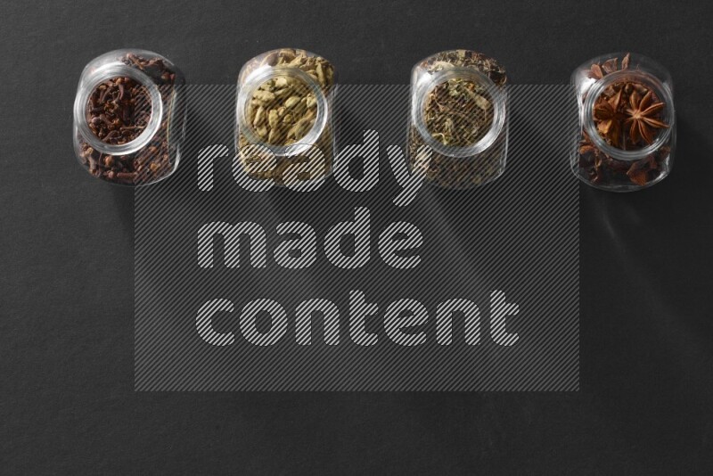 4 glass spice jars full of cloves, cardamom, basil and star anise on black flooring