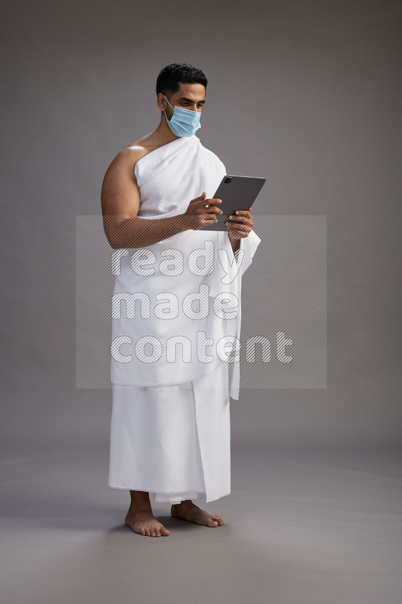 A man wearing Ehram with face mask Standing holding a tablet on gray background
