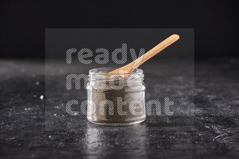 A glass jar full of black pepper powder and a wooden spoon on a textured black flooring