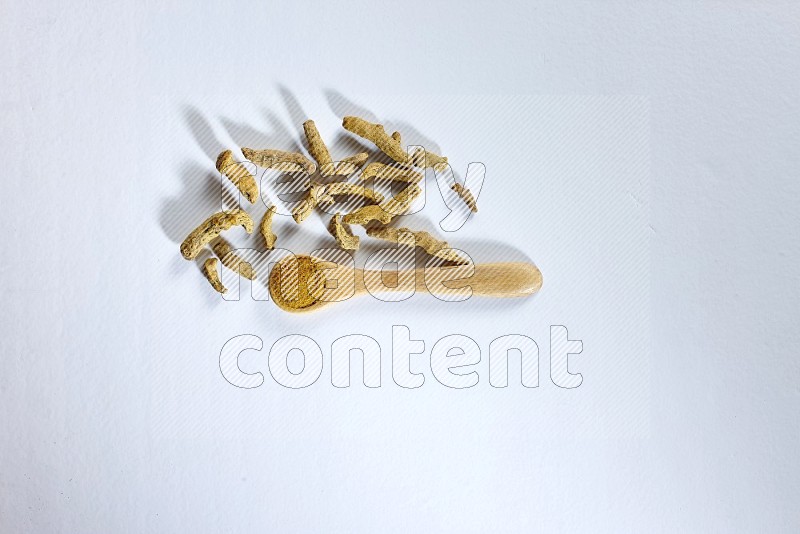 A wooden spoon full of turmeric powder with dried turmeric fingers beside it on white flooring