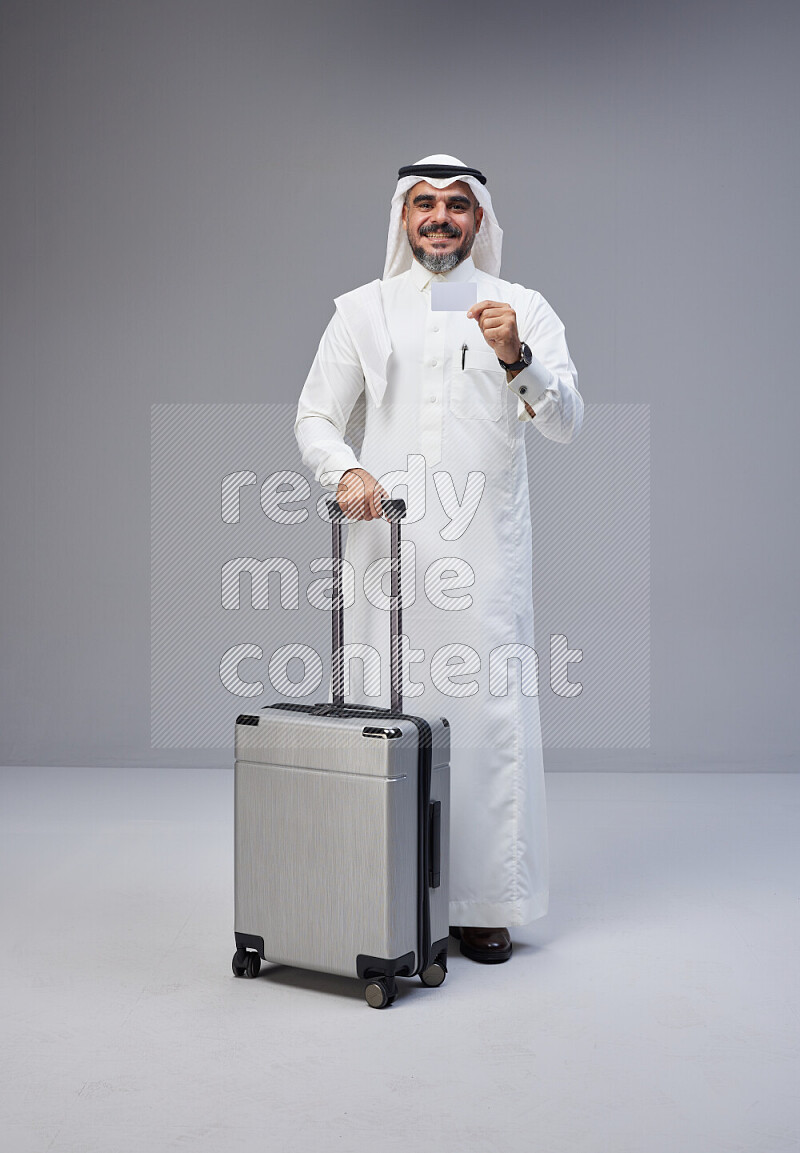 Saudi man wearing Thob and white Shomag standing holding Travel bag and ATM card on Gray background