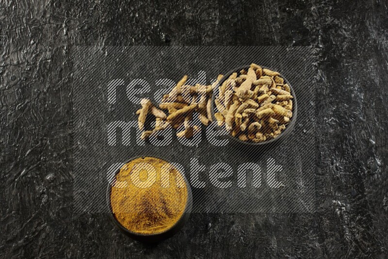 2 pottery black bowls, one full of turmeric powder and the other full of dried turmeric whole fingers on a textured black flooring