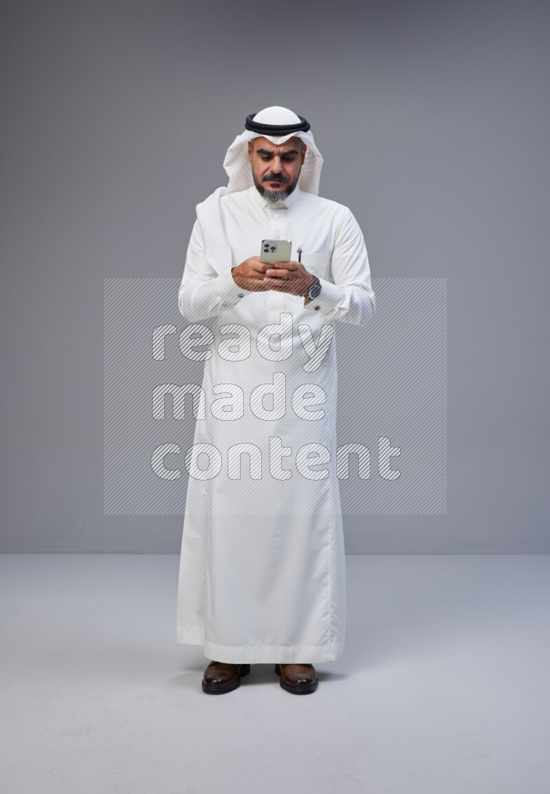 Saudi man Wearing Thob and white Shomag standing texting on phone on Gray background
