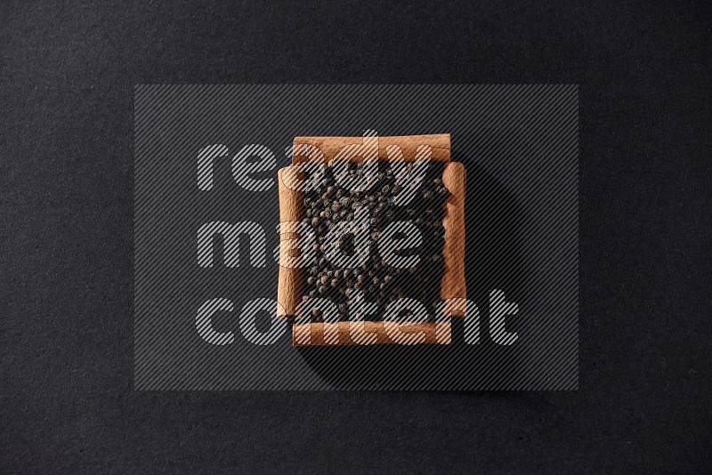 أربعة من أعواد القرفة مكونة شكل مربع بداخلها حبوب الفلفل الأسود علي خلفية سوداء