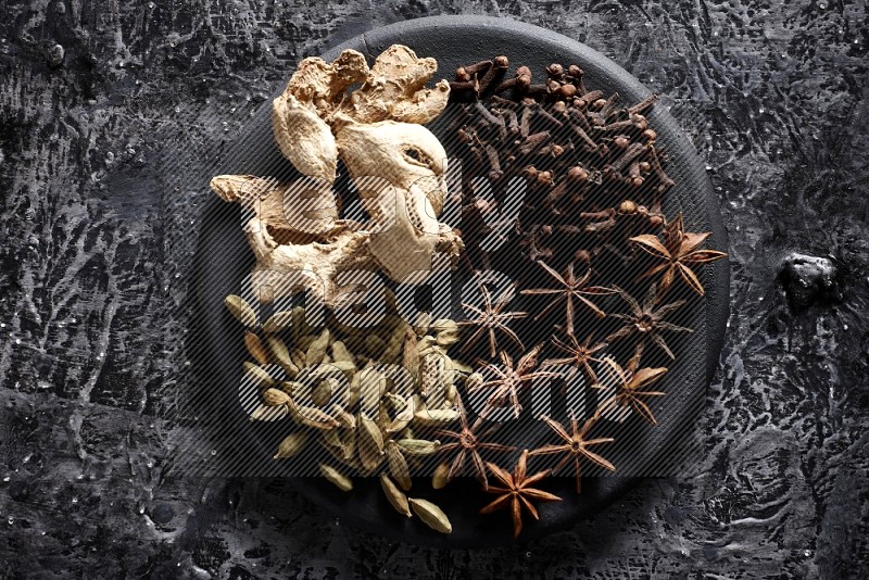 Ginger, cloves, star anise and cardamom on a black plate on textured black background