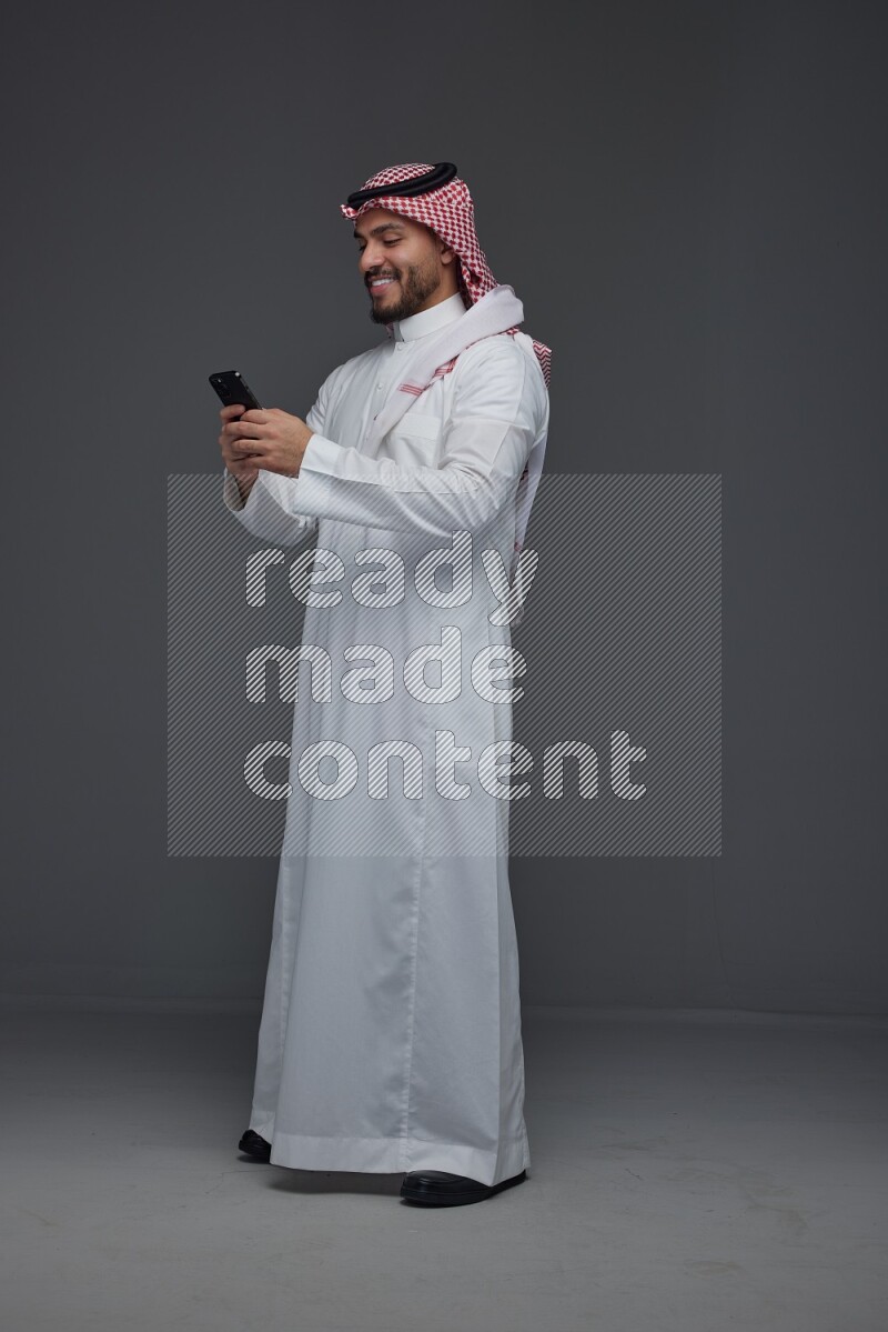 A Saudi man wearing Thobe and Shmagh standing and using his phone eye level on a gray background