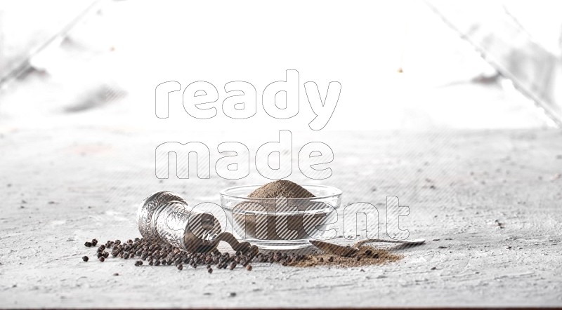 A glass bowl full of black pepper powder with black pepper beads, a turkish metal grinder and a metal spoon on textured white flooring