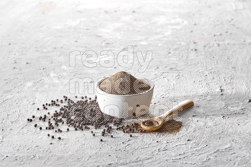 A pottery white bowl full of black pepper powder and wooden spoon full of powder and black pepper beads on a textured white flooring