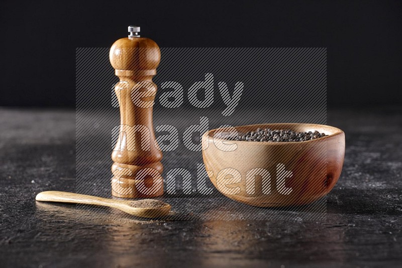 A wooden bowl full of black pepper and a wooden spoon full of black pepper powder and a wooden grinder on a textured black flooring