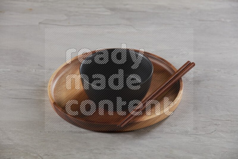 black ceramic round bowl on top of brown wood round plate and wood chopsticks, on grey textured countertop