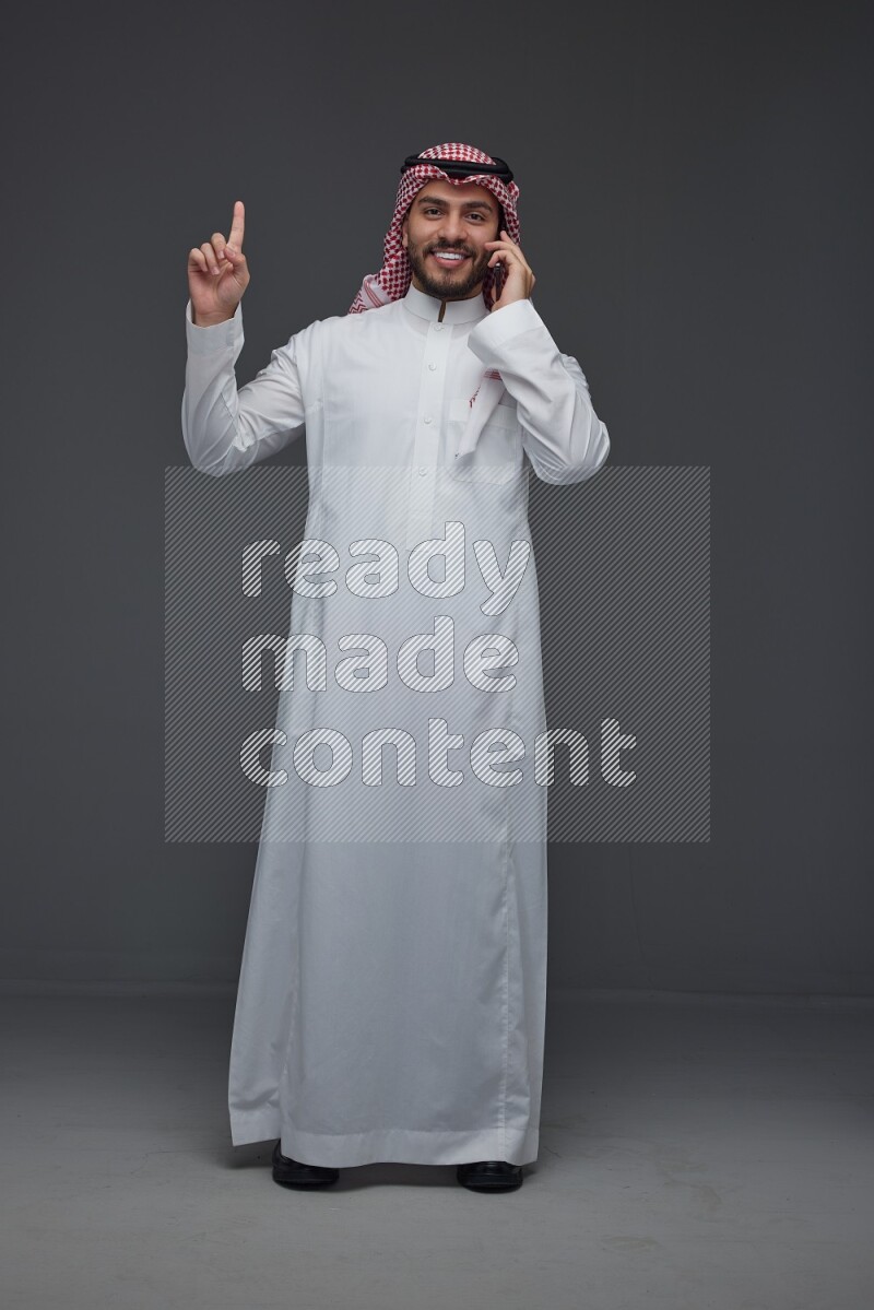 A Saudi man wearing Thobe and Shmagh talking in the phone eye level on a gray background