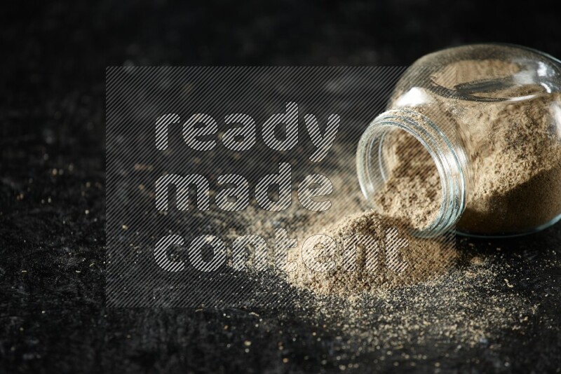 A flipped glass spice jar full of cardamom powder on textured black flooring