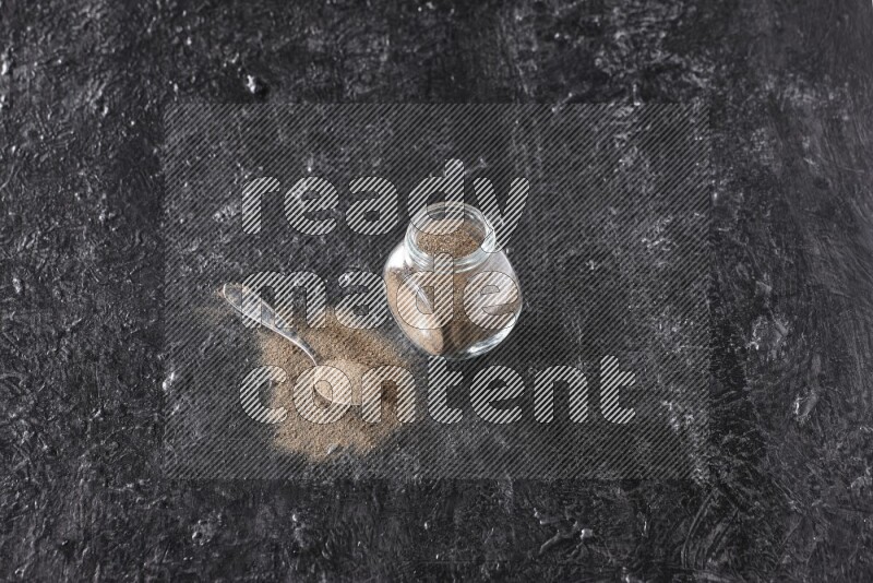 A glass spice jar full of black pepper powder and a metal spoon full of powder on textured black flooring