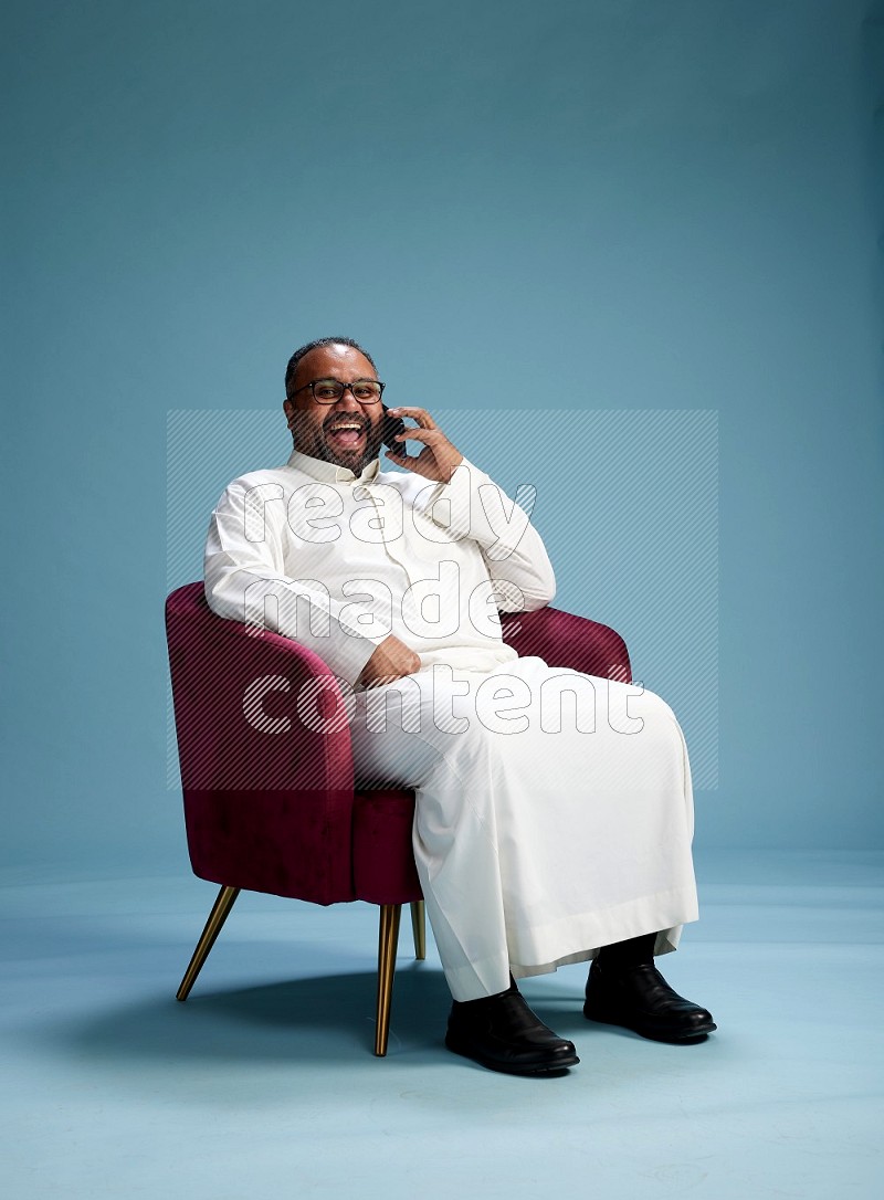 Saudi Man without shimag sitting on chair talking on phone on blue background