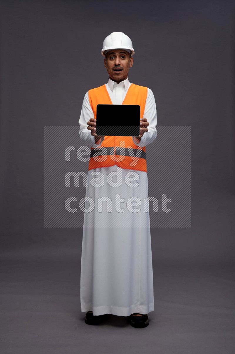 Saudi man wearing thob with engineer vest standing showing tablet to camera on gray background