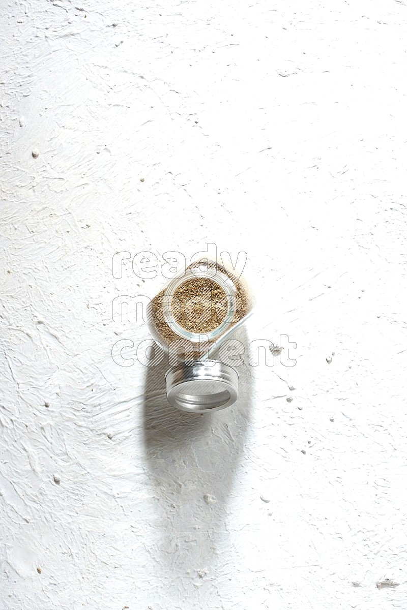 A glass spice jar full of cumin powder on textured white flooring