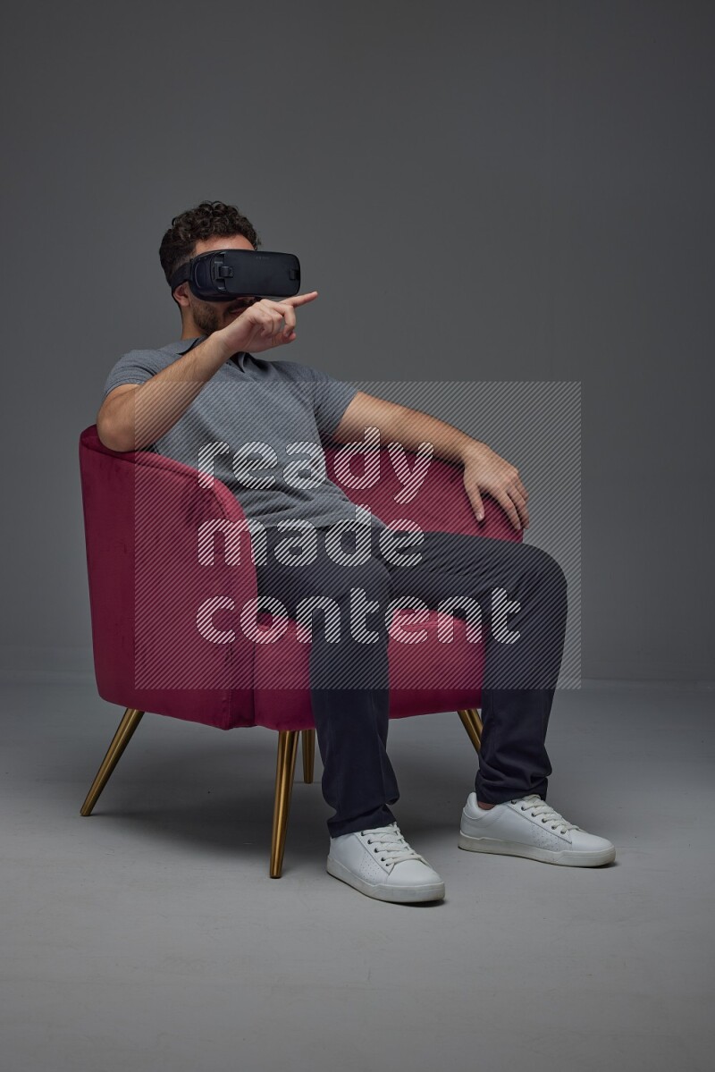 A man wearing casual and wearing VR while sitting on a burgundy chair eye level on a gray background