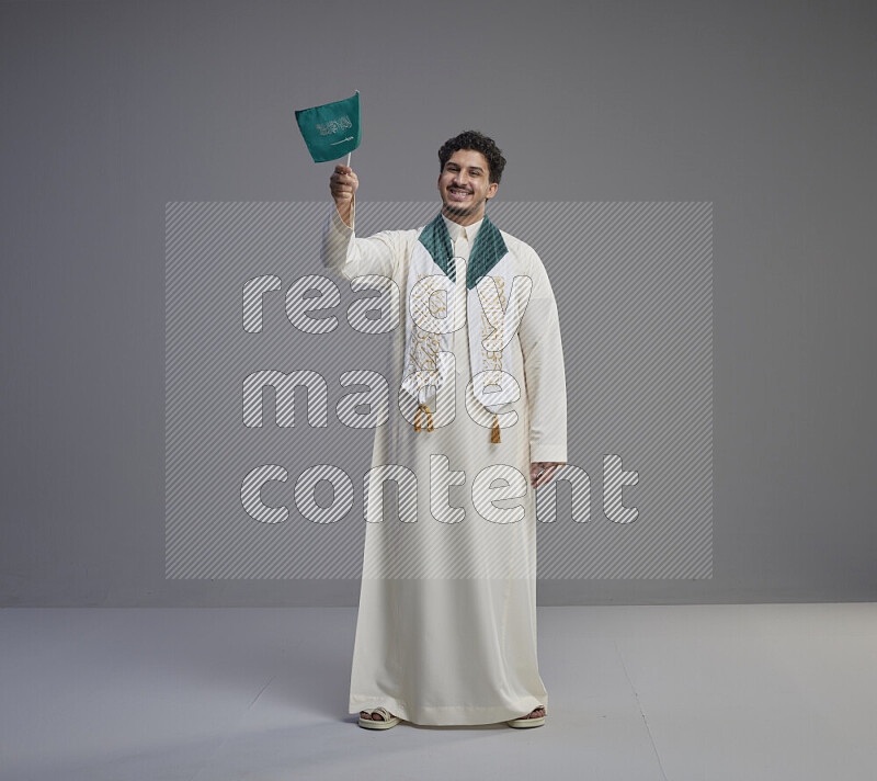 A Saudi man standing wearing thob and saudi flag scarf and holding small saudi flag on gray background