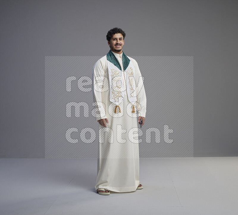 A Saudi man standing wearing thob and Saudi flag scarf on gray background