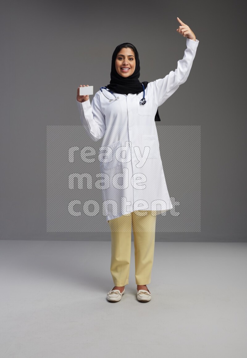 Saudi woman wearing lab coat with stethoscope standing holding ATM card on Gray background