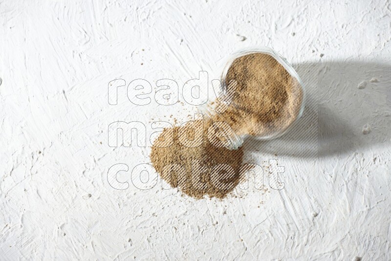 A flipped glass spice jar full of cumin powder and powder spilled out on textured white flooring