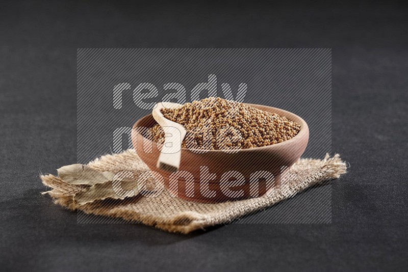 A wooden bowl and spoon full of mustard seeds on a piece of burlap on a black flooring in different angles