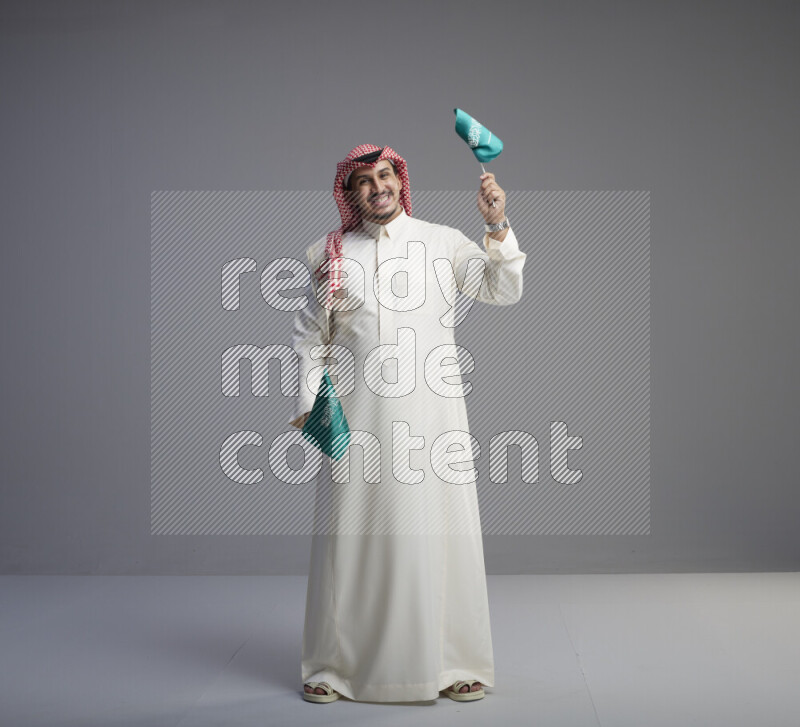 A Saudi man standing wearing thob and red shomag raising small Saudi flag on gray background
