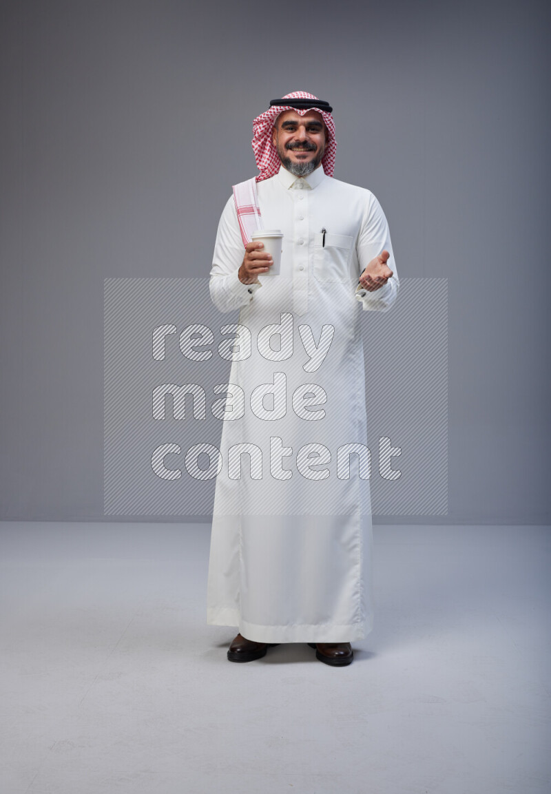 Saudi man Wearing Thob and red Shomag standing holding paper cup on Gray background