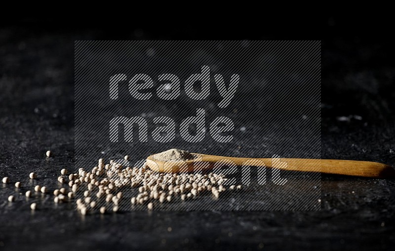 A wooden spoon full of white pepper powder with white pepper beads on textured black flooring